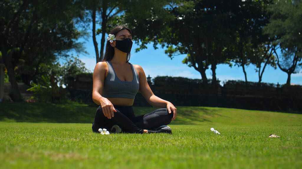 Giovane bruna europea pratica da sola yoga in natura seduta sull'erba verde in posa di loto indossando una maschera protettiva nera sul suo viso - Foto, immagini