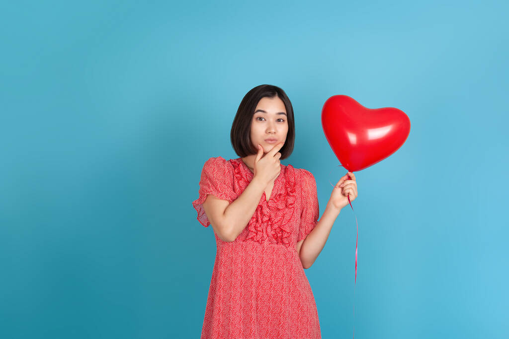 Kırmızı elbiseli, düşünceli genç Asyalı bir kadın kırmızı kalp şeklinde bir balon tutuyor ve mavi arka planda izole edilmiş eliyle çenesini ovuyor. - Fotoğraf, Görsel