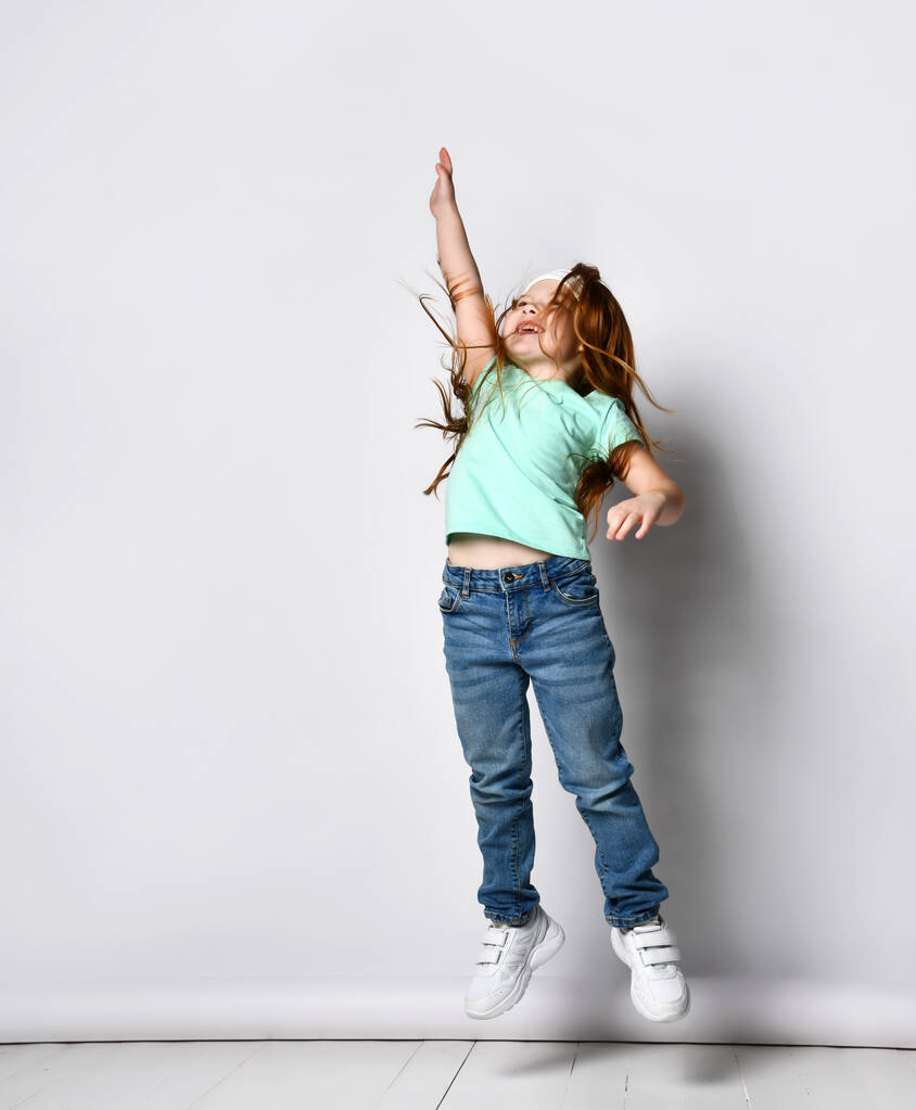 Маленькая счастливая девочка фитнеса, прыгающая с протянутой рукой вверх студии, снятая изолированно на белом копировальном пространстве. Портрет молодого танцора или прыгуна в полный рост. Спорт, тренировки, фитнес и здоровое детство - Фото, изображение