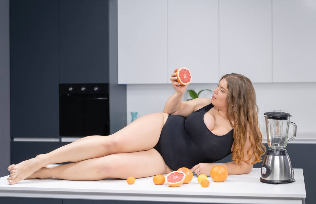Сексуальная толстушка лежит на кухонном столе. Толстая молодая сексуальная пухленькая белая девушка в черном купальнике на современной кухне с фруктами рядом с ней - Фото, изображение
