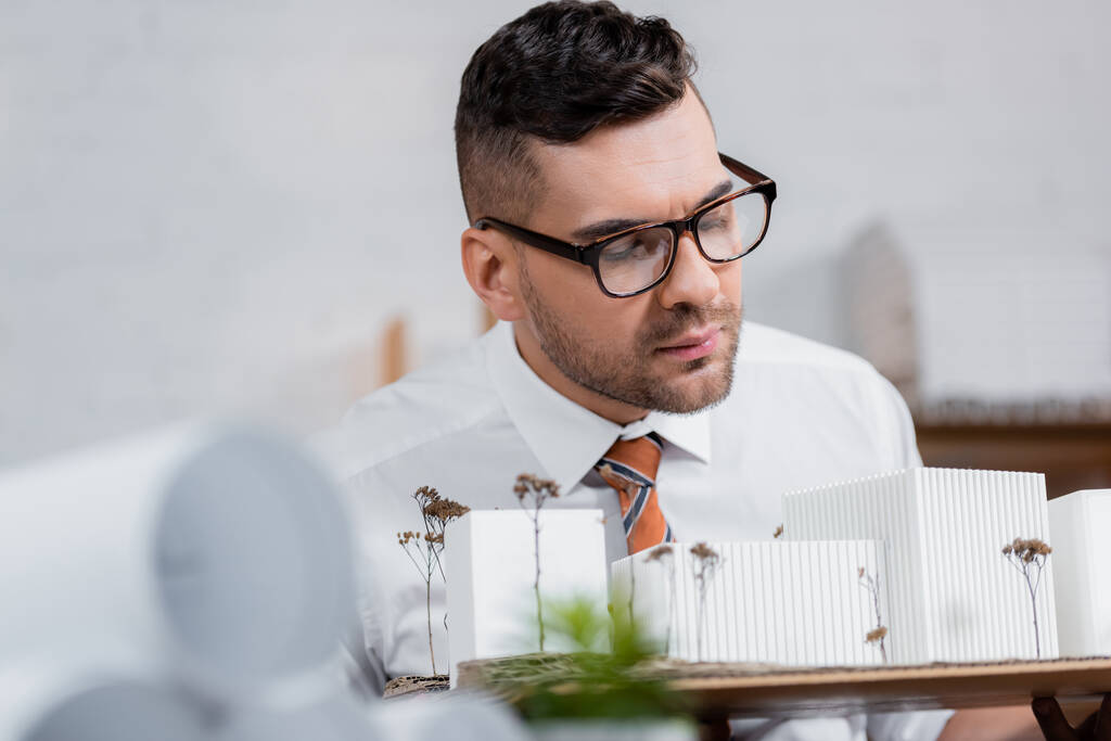 poważny architekt w okularach przyglądający się modelom budynków w biurze, zamazany pierwszy plan - Zdjęcie, obraz
