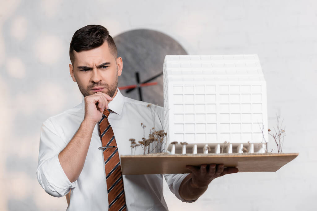 przemyślany architekt z maquette domu trzymając się za rękę w pobliżu podbródka w biurze - Zdjęcie, obraz