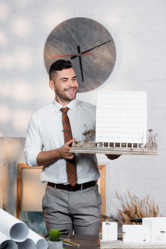 arquitecto positivo sosteniendo la maqueta del edificio mientras está parado en el lugar de trabajo - Foto, imagen