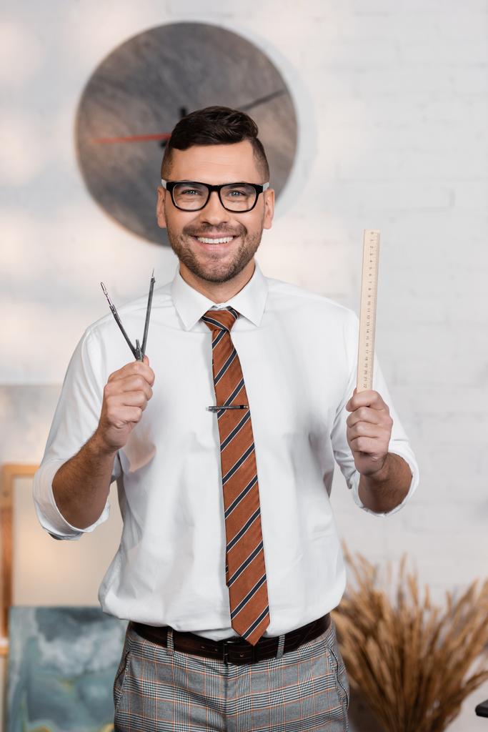 cheerful architect in eyeglasses holding ruler and divider while smiling at camera - Photo, Image