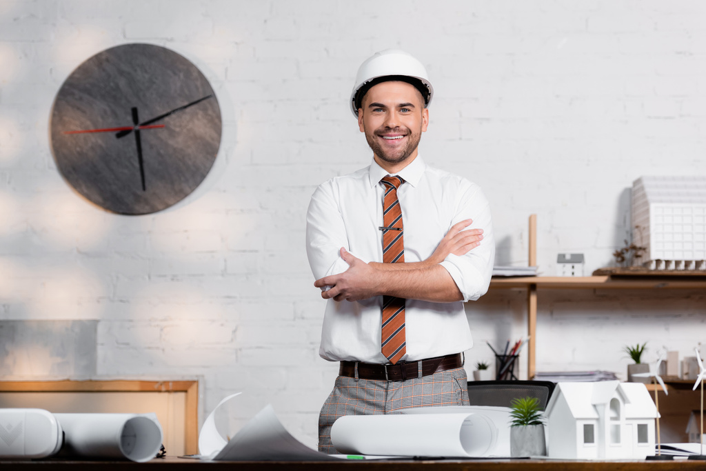 улыбающийся архитектор в шлеме, стоящий со скрещенными руками возле чертежей и модели дома - Фото, изображение
