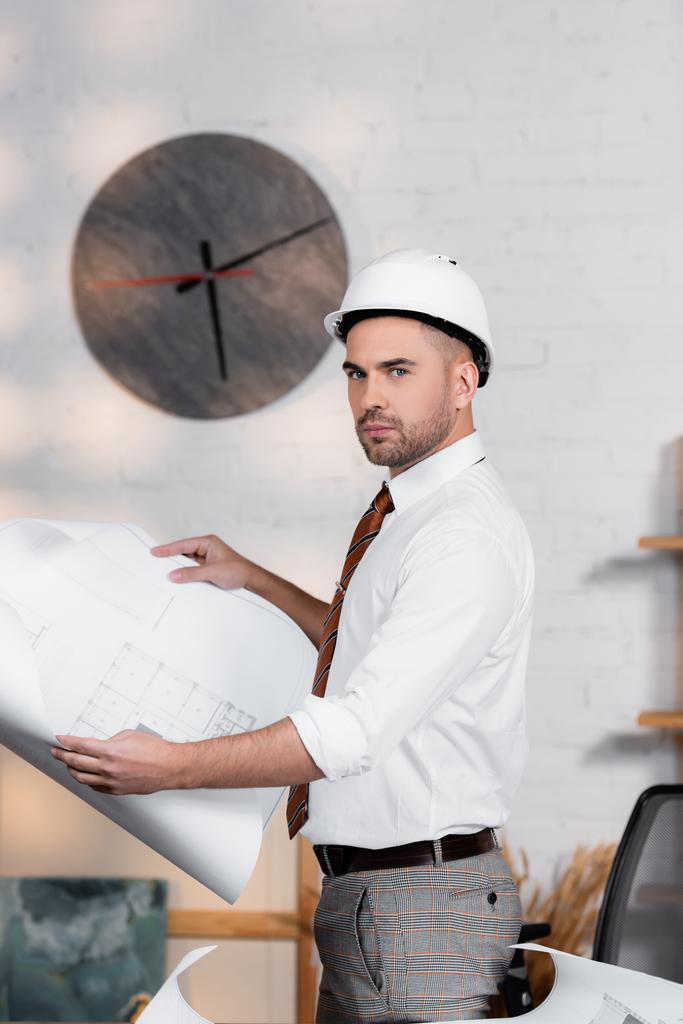 seriózní architekt v helmě drží plán a dívá se do kamery - Fotografie, Obrázek