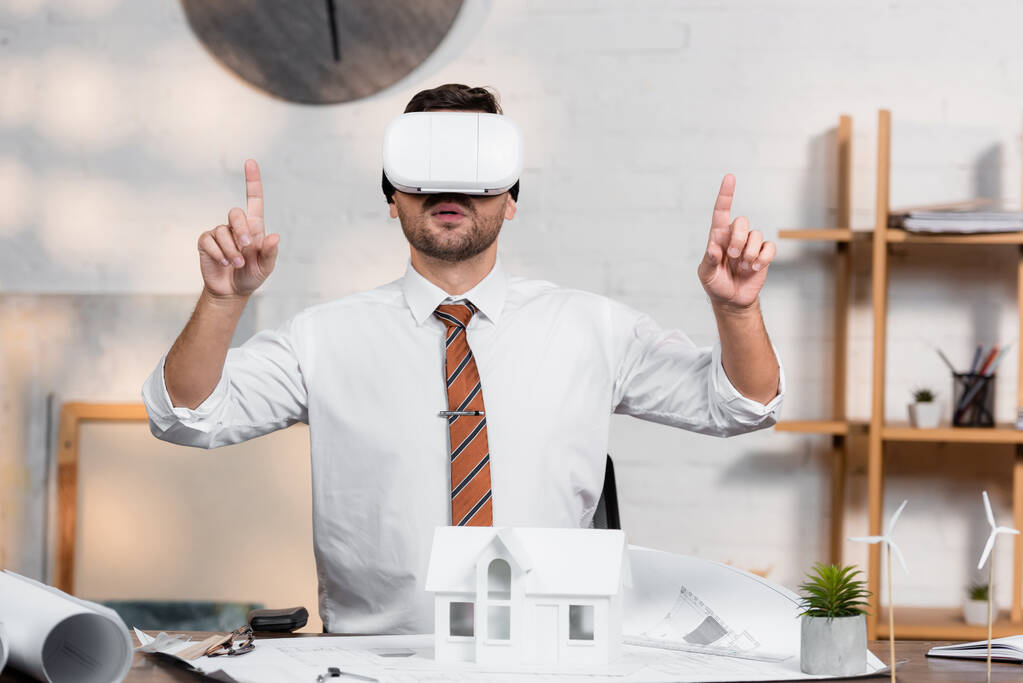 VR kulaklıklı mimar ev modelinin yanındaki iş yerinde otururken parmaklarıyla işaret ediyor - Fotoğraf, Görsel