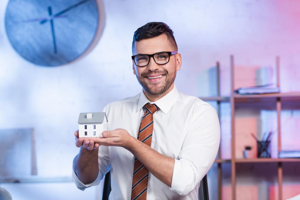 весёлый архитектор в очках держит модель дома и улыбается в камеру - Фото, изображение