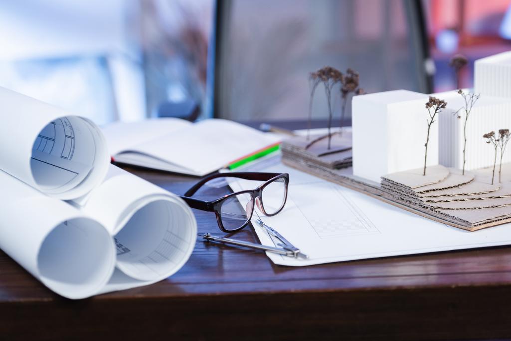 selective focus of eyeglasses near house models and blueprints on desk in office, blurred background - Photo, Image