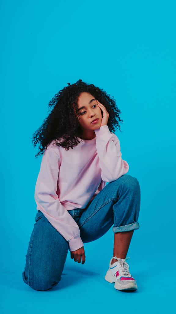 A beautiful curly haired girl posing in a pink sweater and jeans on a blue background - Photo, Image