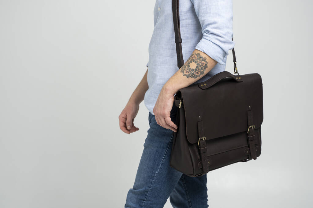 Brown mens shoulder leather bag for a documents and laptop on the shoulders of a man in a blue shirt and jeans with a white background. Satchel, mens leather handmade briefcase. - Photo, Image