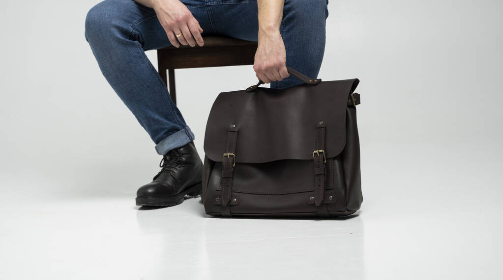 Άνδρας με μπλε τζιν και μαύρες μπότες κάθεται σε μια καρέκλα με μια καφέ mens τσάντα δερματίνης ώμου για ένα έγγραφο και φορητό υπολογιστή σε ένα λευκό πάτωμα. Δερμάτινη τσάντα mens, τσάντες messenger, χειροποίητος χαρτοφύλακας. - Φωτογραφία, εικόνα