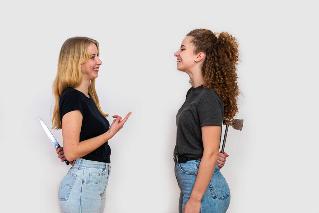 two smiling women standing face to face and hiding knife and axe behind back, isolated on white background, the concept of hidden hatred and hypocrisy - Photo, Image