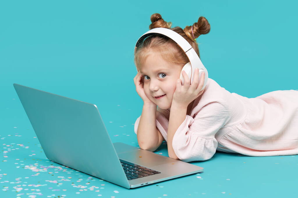 Älykäs lapsi tyttö langattomissa kuulokkeissa syaanitaustalla kannettavan tietokoneen avulla. Koulutus, verkko-opiskelu, televisio, etäopetus. Peruskoulun oppilas karanteenin aikana. Opetuspelit lapsille tietokoneella - Valokuva, kuva