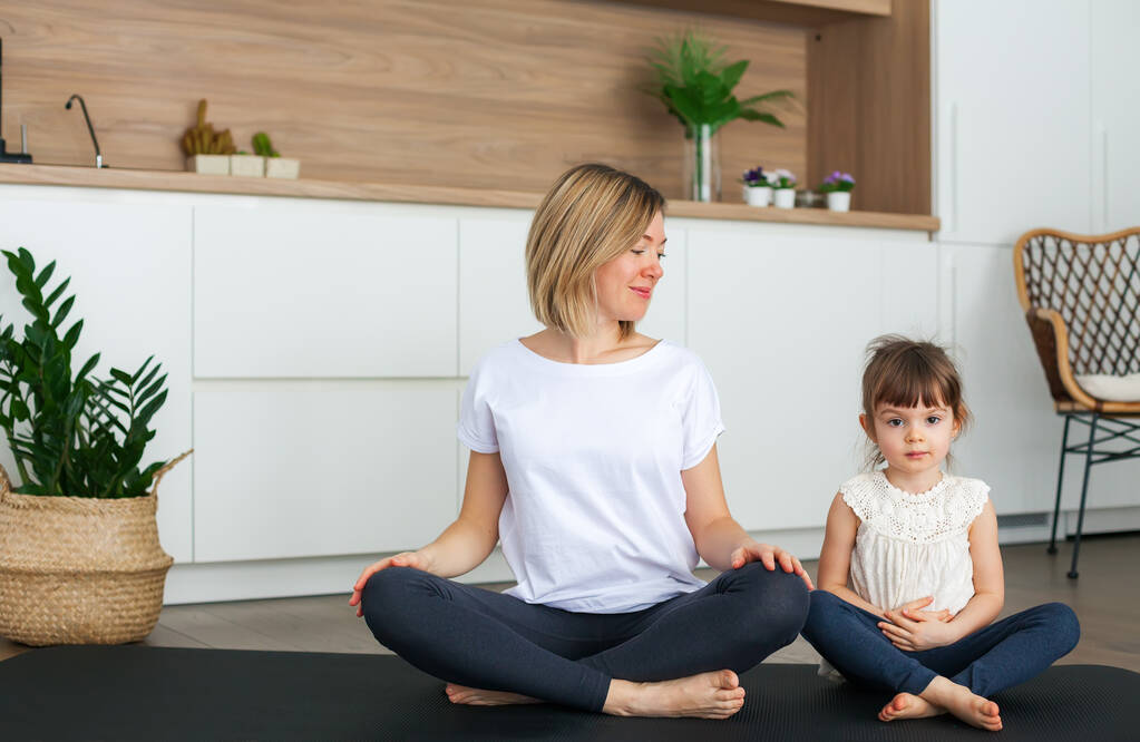 Kadın ve küçük tatlı kızı evde yoga yaparken lotus pozisyonunda oturuyorlar. - Fotoğraf, Görsel