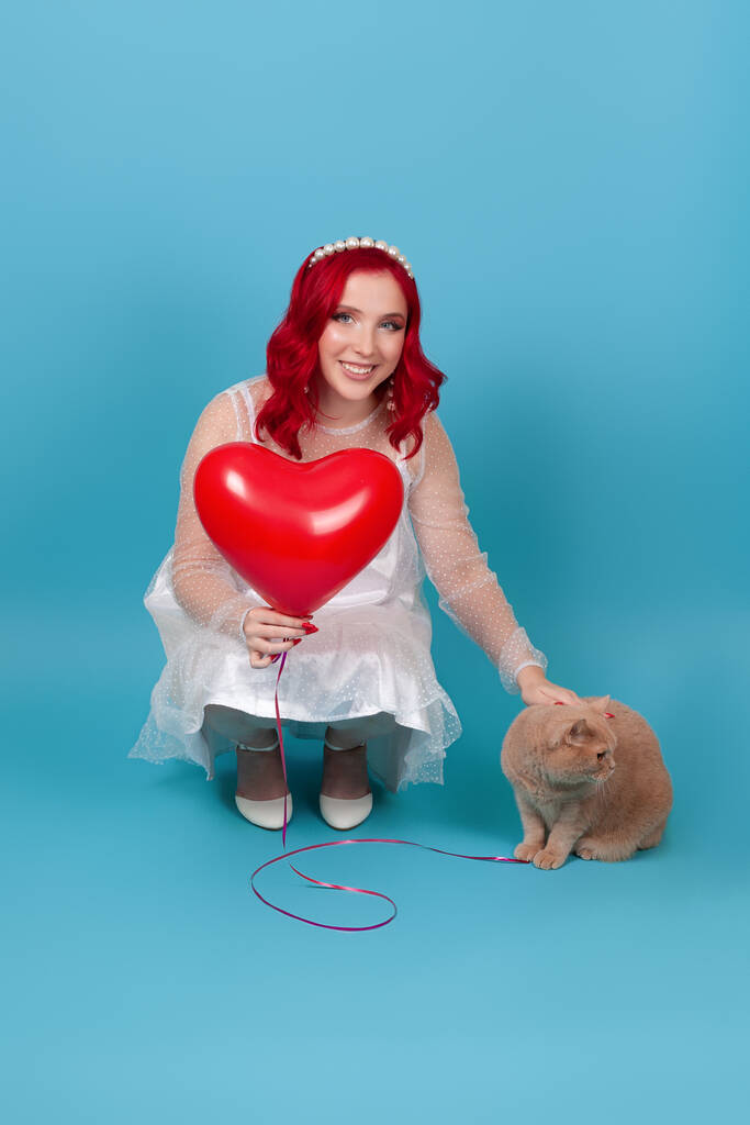 Beyaz elbiseli, gülümseyen genç bir kadın kalp şeklinde bir balon tutuyor, çömeliyor ve mavi arka planda izole edilmiş kırmızı bir İngiliz kedisini sıkıyor. - Fotoğraf, Görsel