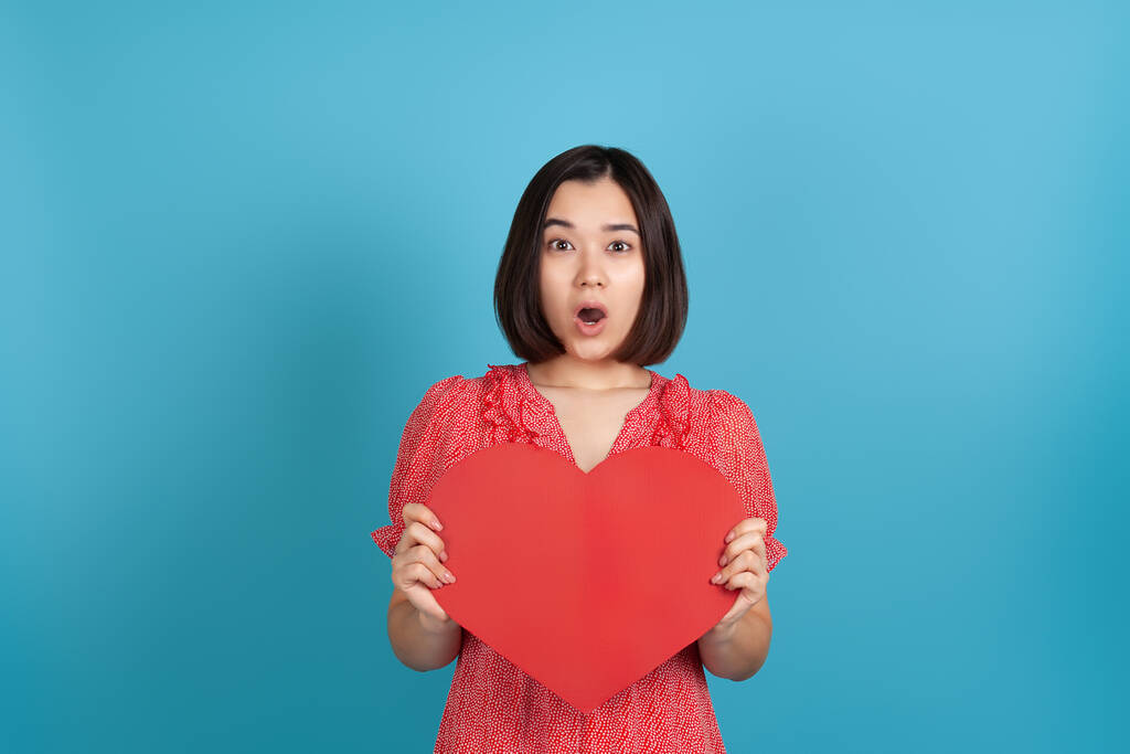 Крупный план удивлен, взволнован, впечатлен молодой азиатской женщины с открытым ртом в красном платье проведение большой красной бумаги сердце изолированы на синем фоне - Фото, изображение