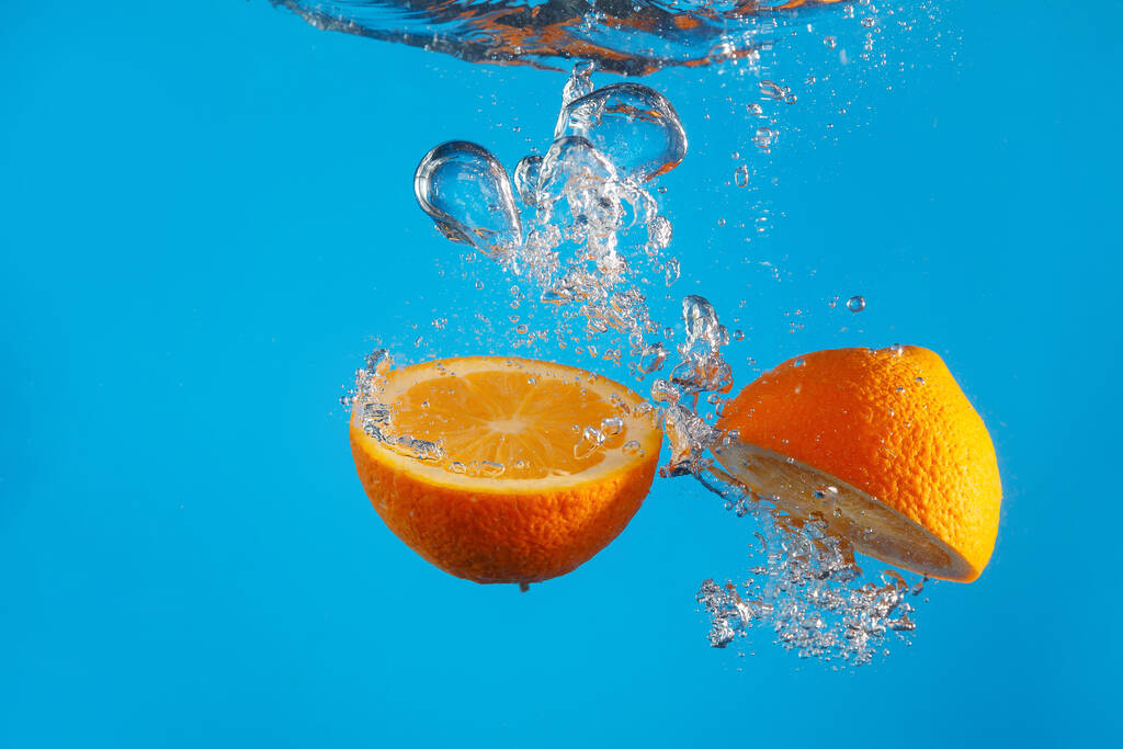 Two halves of an orange falls into the water with a splash on a blue background - Photo, Image