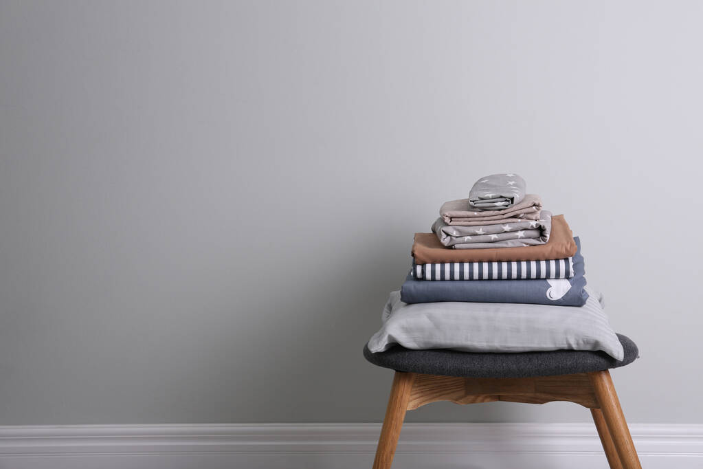 Стопка чистого постельного белья и подушки на табуретке возле светло-серой стены. Пространство для текста - Фото, изображение
