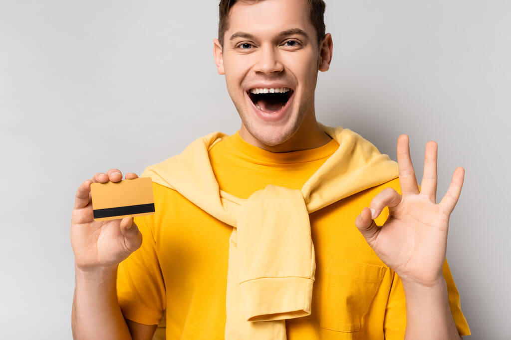 Homme joyeux avec carte de crédit montrant geste correct sur fond gris - Photo, image