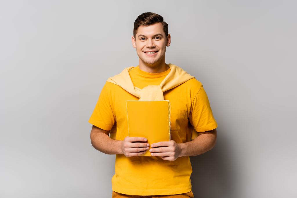 Χαμογελώντας άνθρωπος κρατώντας κίτρινο σημειωματάριο σε γκρι φόντο - Φωτογραφία, εικόνα