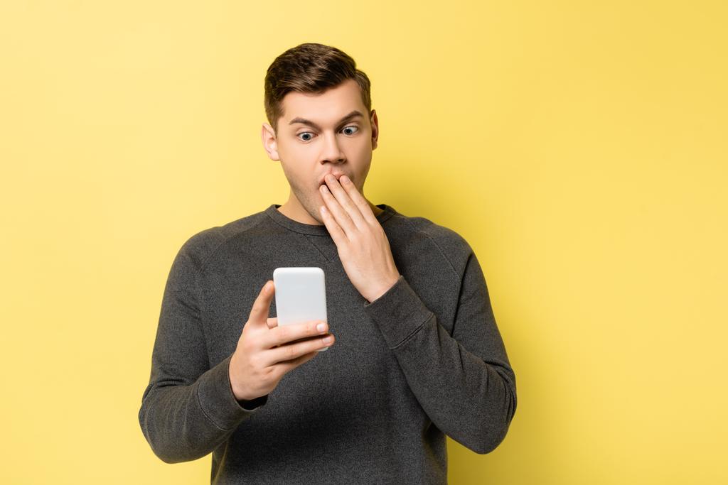 Niesamowity mężczyzna zakrywający usta trzymając smartfon na żółtym tle - Zdjęcie, obraz