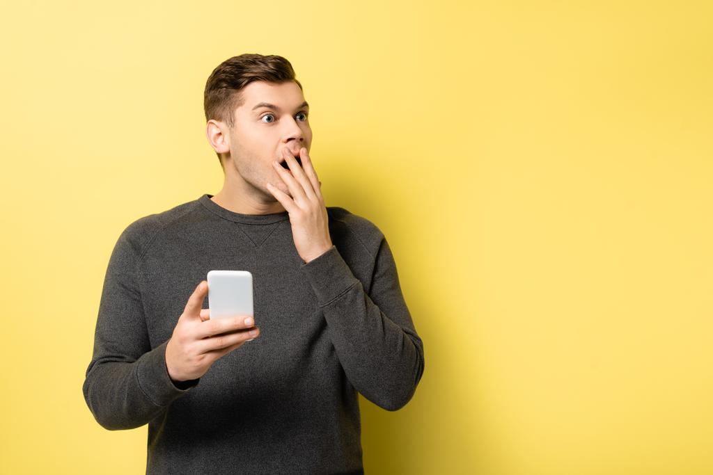 Podekscytowany mężczyzna z ręką w pobliżu ust trzyma telefon komórkowy na żółtym tle - Zdjęcie, obraz