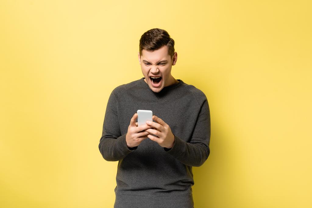 Εκνευρισμένος άντρας ουρλιάζει ενώ χρησιμοποιεί κινητό σε κίτρινο φόντο. - Φωτογραφία, εικόνα