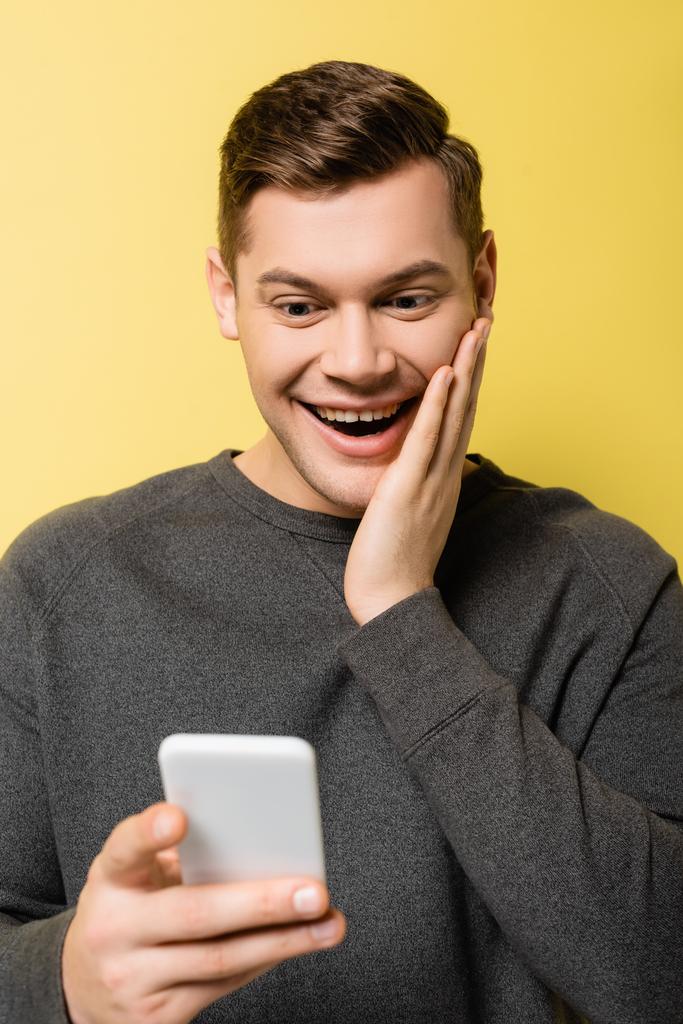 Hombre feliz con la mano cerca de la mejilla sosteniendo el teléfono inteligente en primer plano borroso sobre fondo amarillo - Foto, Imagen