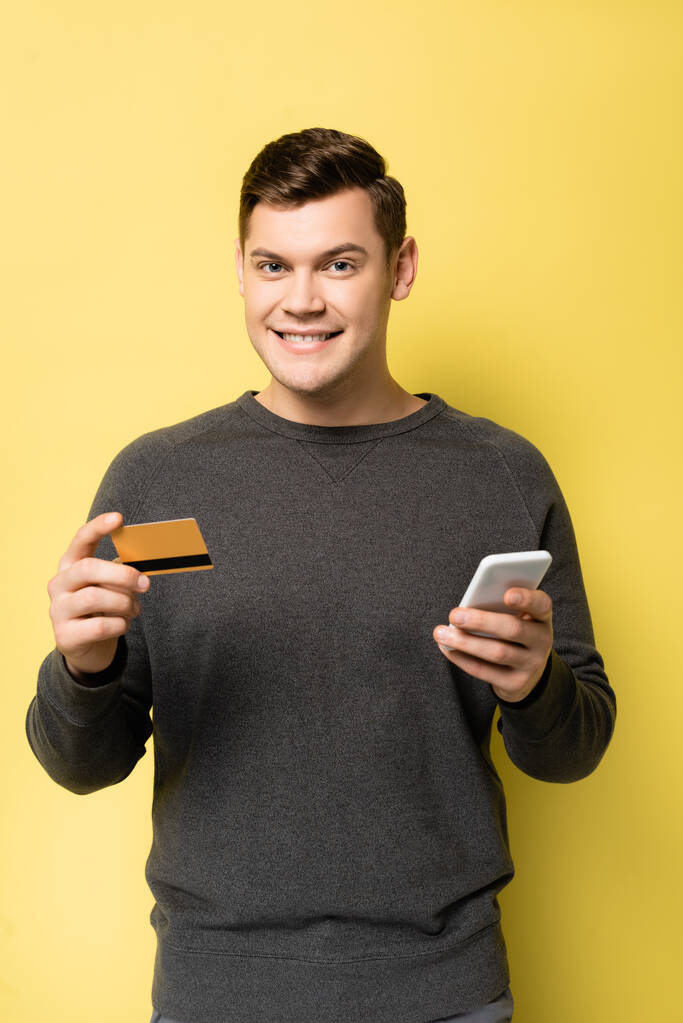 黄色い背景のカメラを見ながらクレジットカードとスマートフォンを持った笑顔の男 - 写真・画像