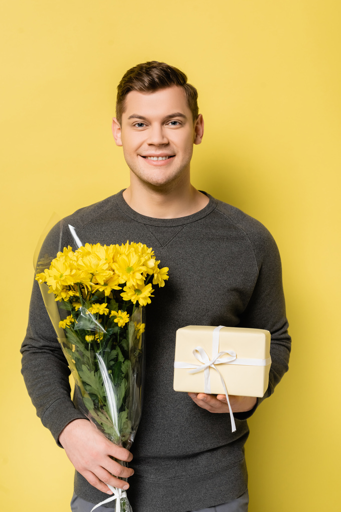 Νεαρός άνδρας χαμογελά στην κάμερα κρατώντας κουτί δώρου και μπουκέτο σε κίτρινο φόντο - Φωτογραφία, εικόνα
