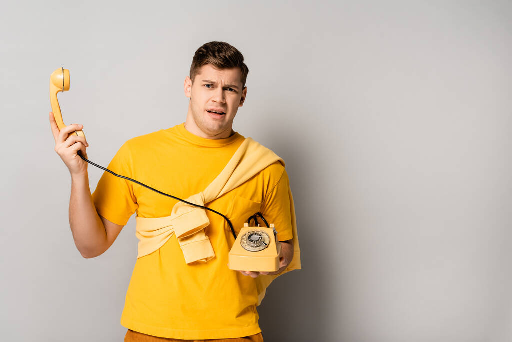 Embarrassed man holding yellow retro telephone on grey background - Photo, Image