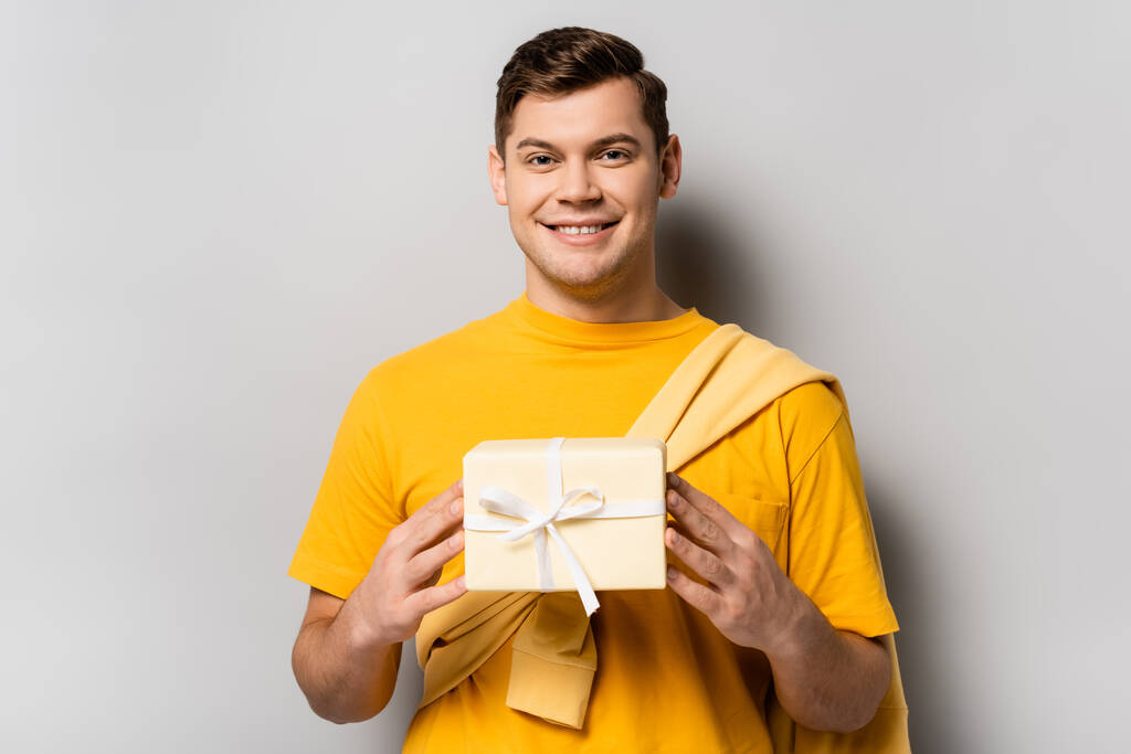 Heureux homme brune tenant cadeau avec ruban sur fond gris - Photo, image