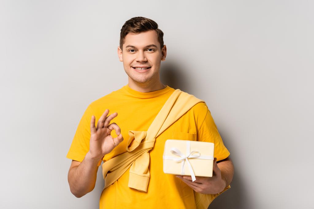 Молодой человек с подарочной коробкой показывает нормальный жест на сером фоне - Фото, изображение