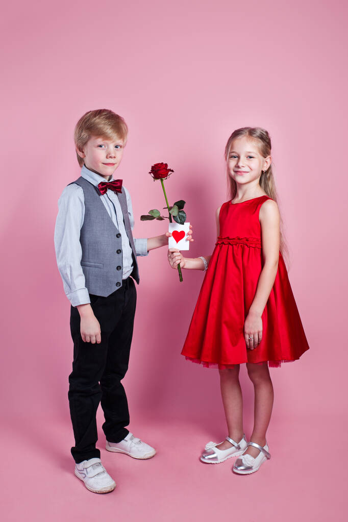 Niespodzianka na Walentynki. Mały chłopiec w miłości dając słodkie dziewczyny czerwona róża na różowym tle , - Zdjęcie, obraz