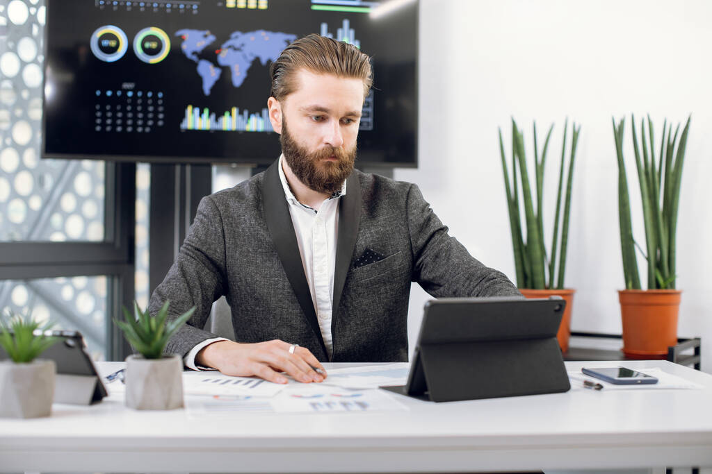 Portret van goed uitziende, geconcentreerde mannelijke kantoormanager met baard, die bedrijfskleding draagt, werkt bij i-pad tablet en financiële gegevens analyseert in projectdocumenten - Foto, afbeelding