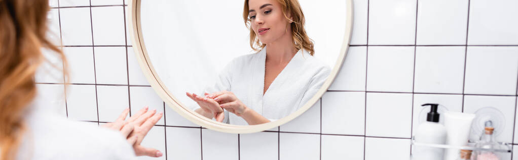 женщина наносит крем для рук возле зеркала в ванной комнате, баннер - Фото, изображение