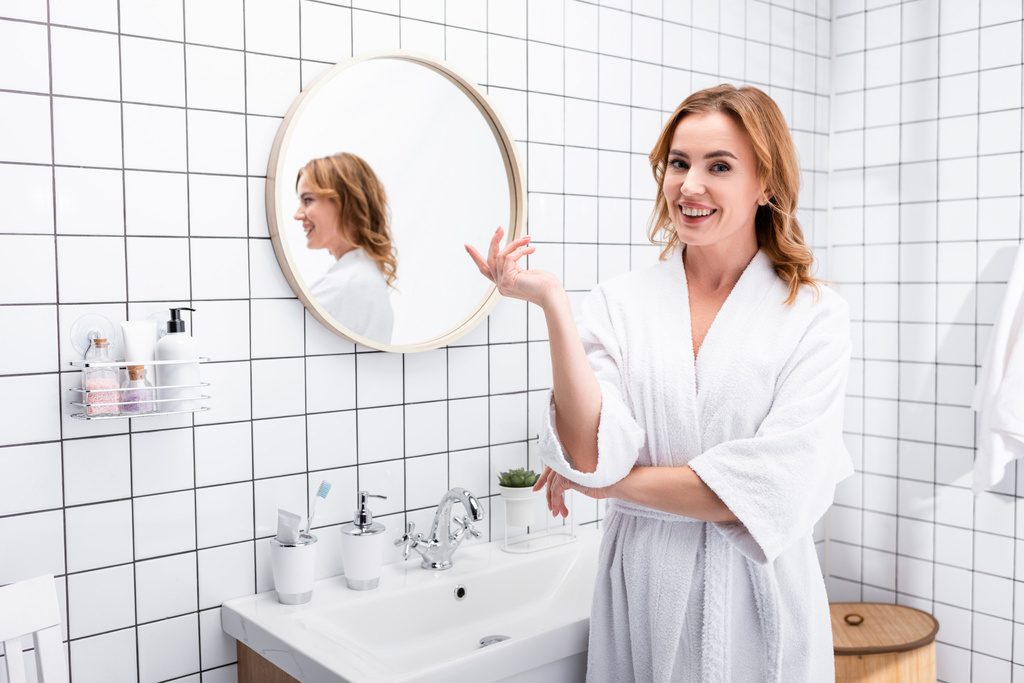 веселая женщина в халате улыбается во время жеста в ванной комнате  - Фото, изображение
