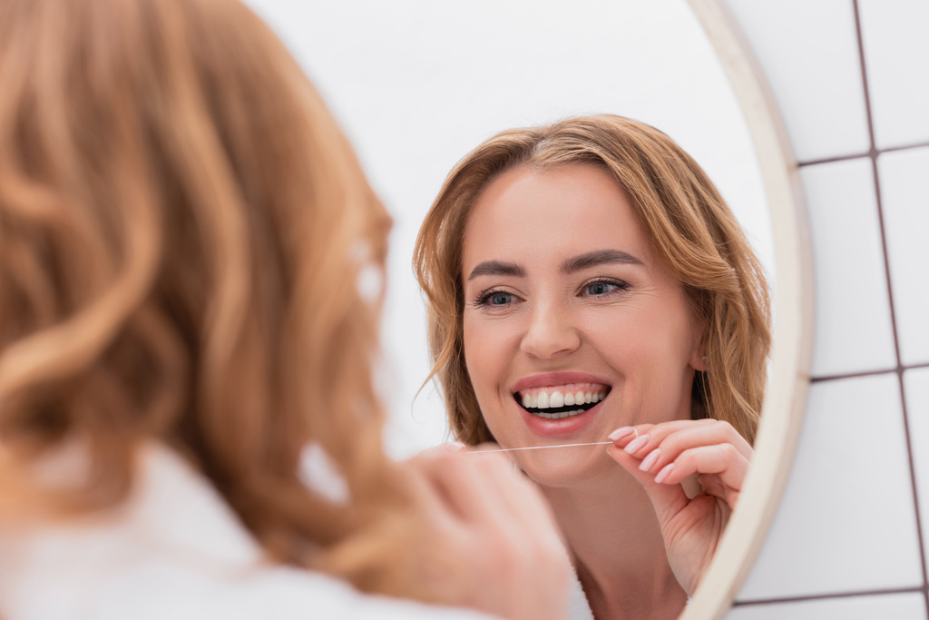 Spiegelbild einer fröhlichen Frau, die in den Spiegel schaut und Zahnseide in der Hand hält - Foto, Bild