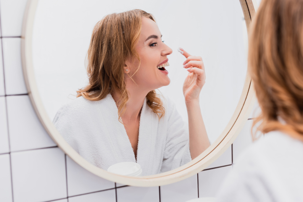 улыбающаяся женщина, держащая банку и нанося крем для лица, глядя в зеркало  - Фото, изображение