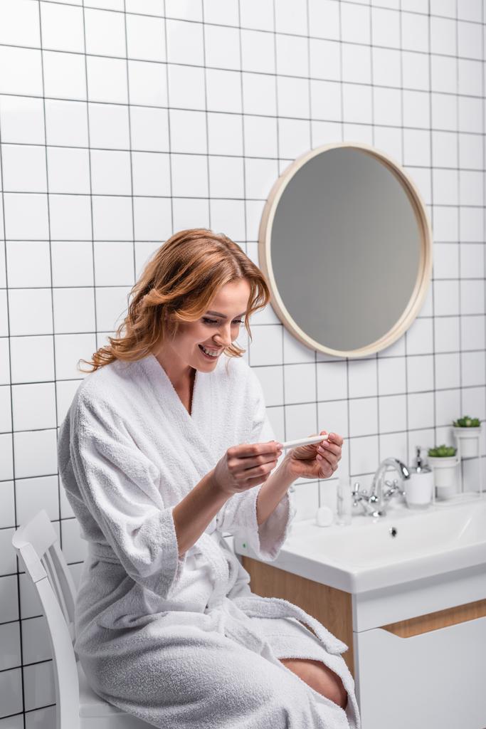 Beyaz bornozlu mutlu kadın banyoda hamilelik testine bakıyor. - Fotoğraf, Görsel