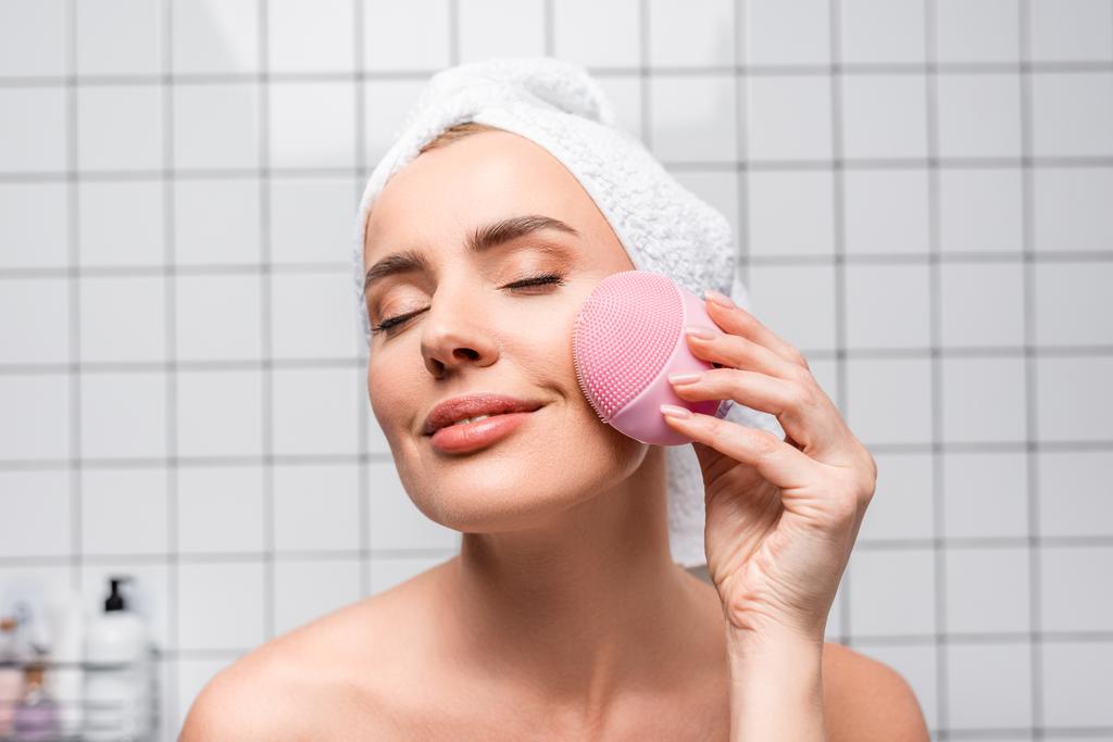 mujer alegre en toalla en la cabeza y los ojos cerrados sosteniendo cepillo de silicona limpiadora en el baño - Foto, imagen