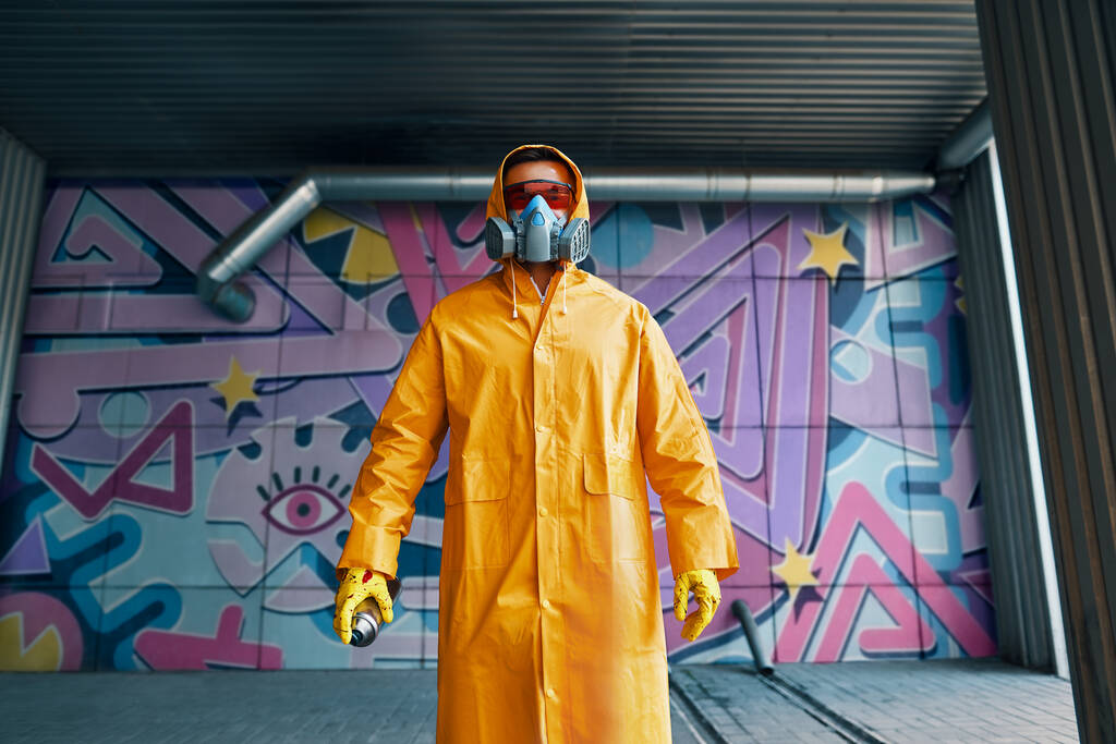 Γκράφιτι ζωγράφος με μάσκα αναπνοής στέκεται κοντά στον τοίχο με τους πίνακές του - Φωτογραφία, εικόνα