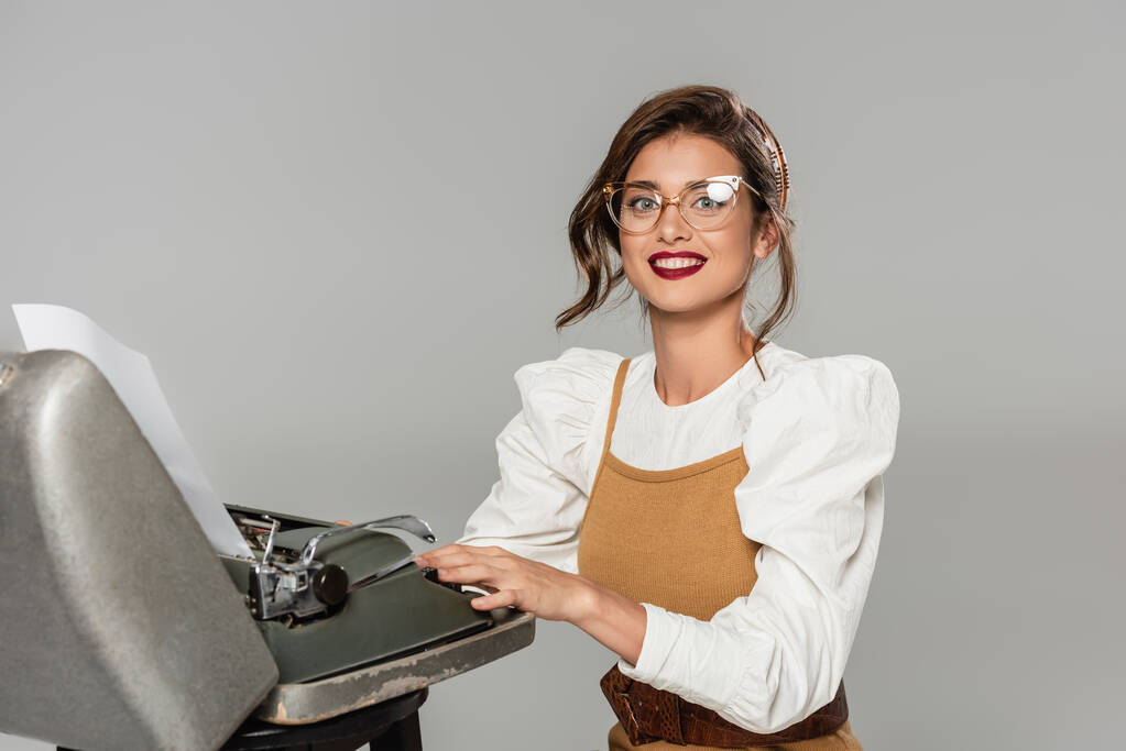 fröhliche Sekretärin tippt auf Retro-Schreibmaschine und lächelt in die Kamera isoliert auf grau - Foto, Bild