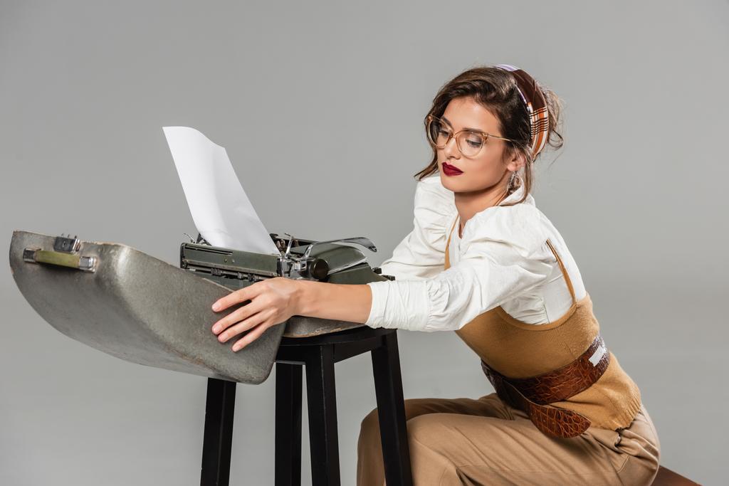 стильная секретарша, открывающая корпус винтажной пишущей машинки, изолированной на сером - Фото, изображение