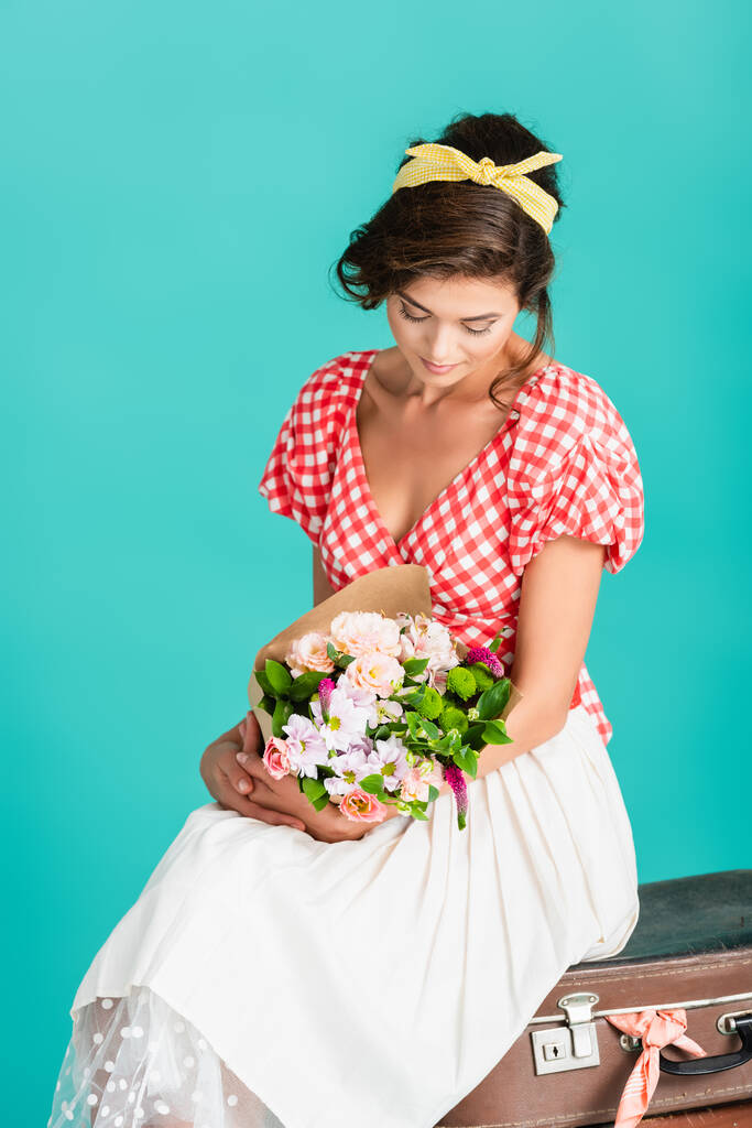 elegante pin-up mujer sosteniendo flores mientras está sentado en la maleta vintage aislado en turquesa - Foto, imagen