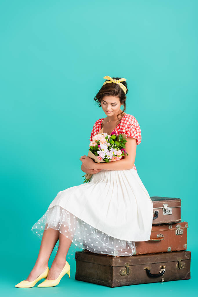 молодая женщина в ретро-одежде сидит на винтажных чемоданах с букетом на бирюзовом - Фото, изображение