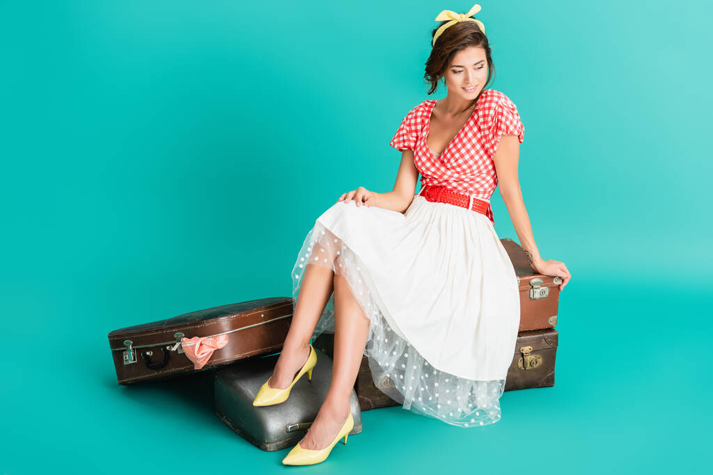 femme heureuse en vêtements rétro assis sur des valises vintage près de la machine à écrire au cas où sur turquoise - Photo, image