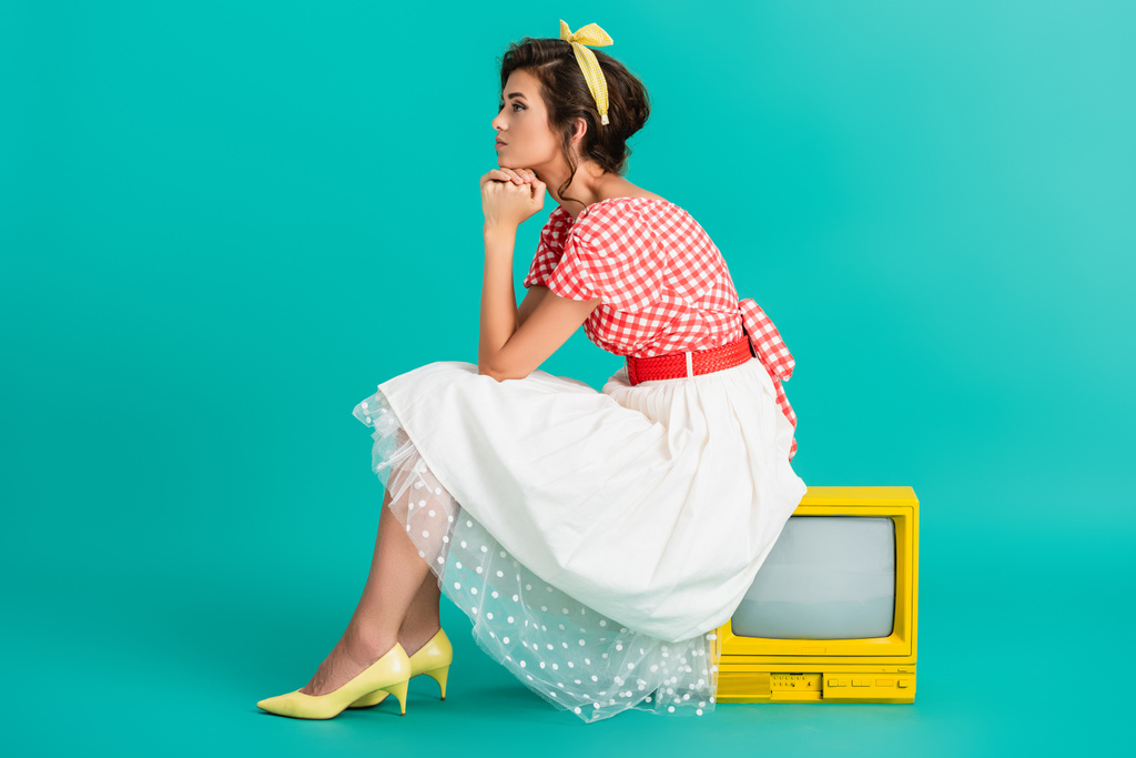 вид сбоку мечтательной женщины, сидящей на желтом винтажном телевизоре, установленном на бирюзе - Фото, изображение