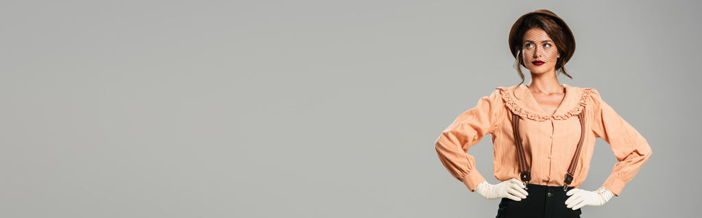 красивая молодая женщина в модной ретро-одежде и перчатках позирует с руками на бедрах изолированных на сером, баннер - Фото, изображение